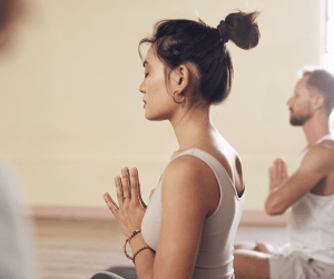 A woman in the forefront of the image meditating in a class with a male also meditating to the back right of the image 
